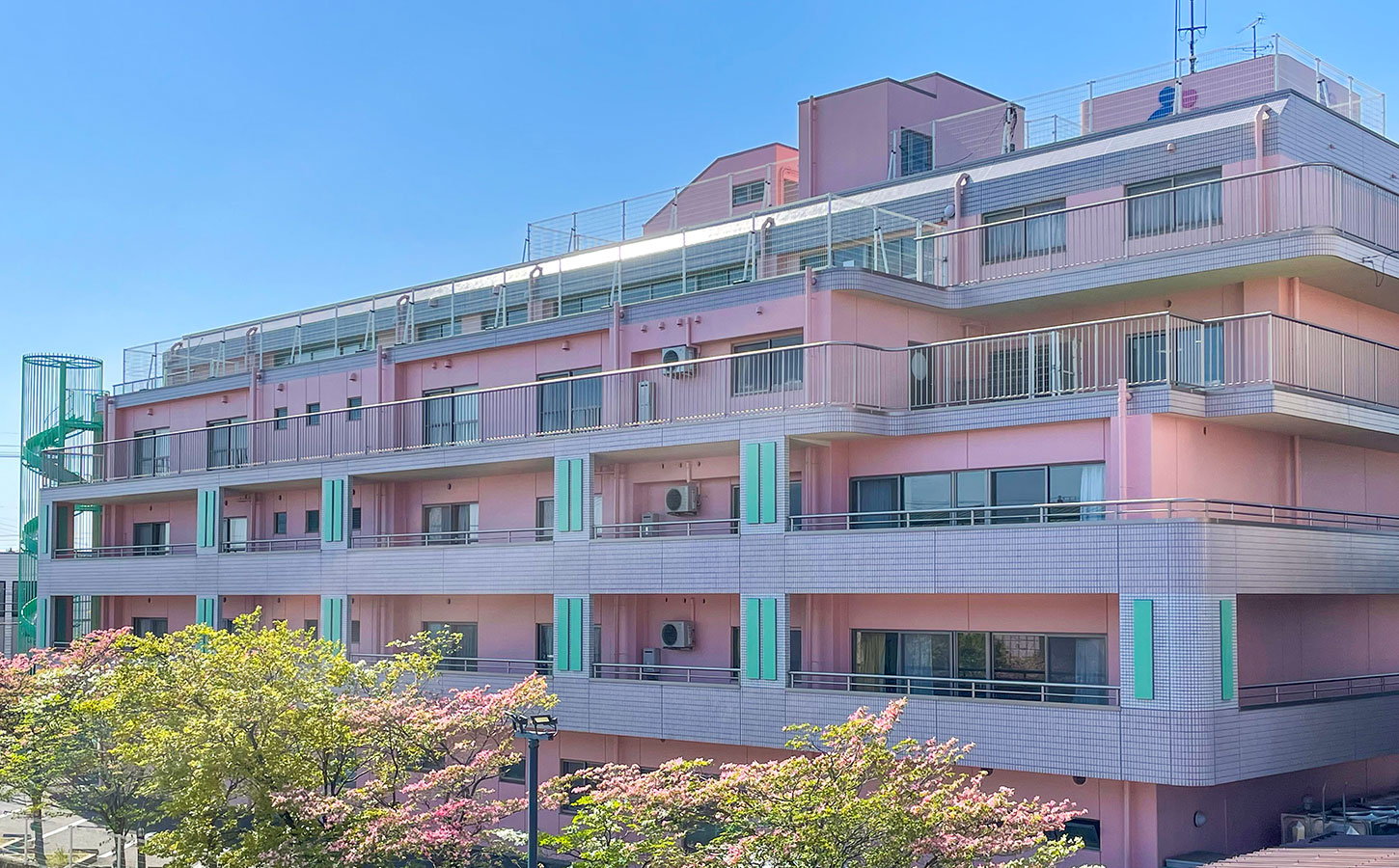 ピンク色の外壁を持つ介護施設の建物外観。複数階建てで緑のアクセントが特徴。
